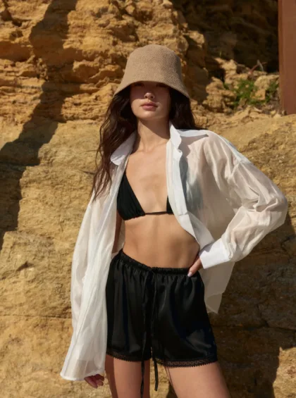 Woman in a black bikini top and sheer white shirt stands against rocky beige cliffs, wearing a beige bucket hat.