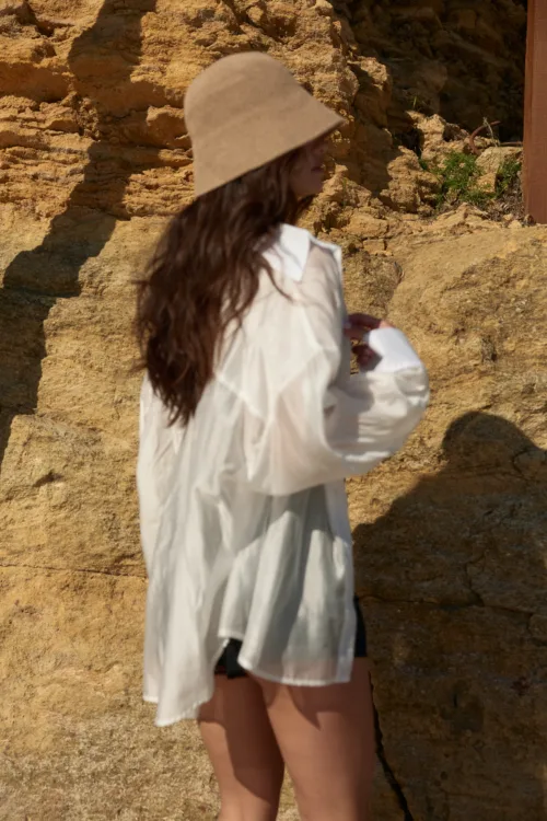 Person wearing a wide-brim beige hat and white shirt standing against a rocky beige wall, facing sideways.