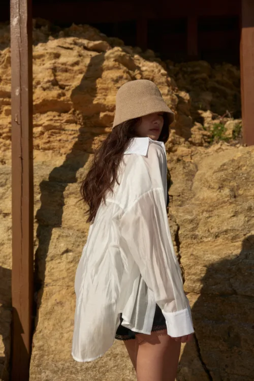 Woman in a beige knit hat and white oversized shirt standing outdoors against rocky terrain, partial side view.