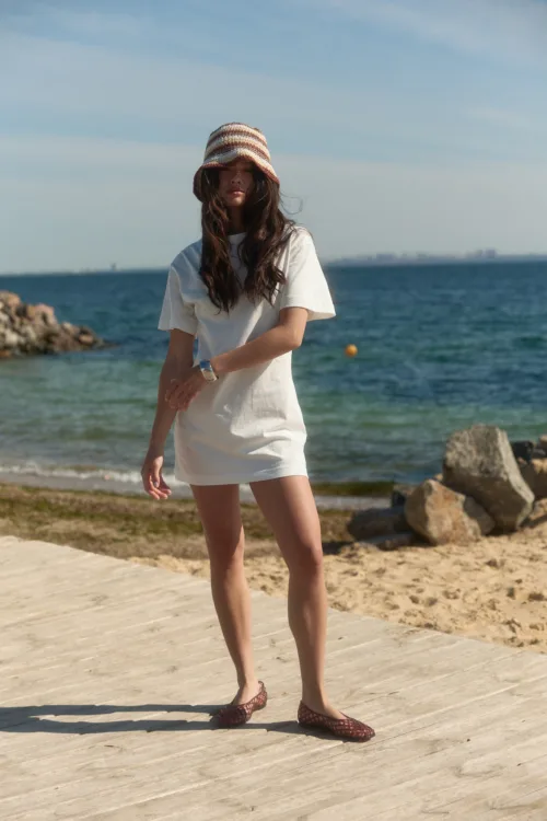 Woman in a white dress and striped sunhat walking on a wooden boardwalk by the sea, with calm blue water in the background.