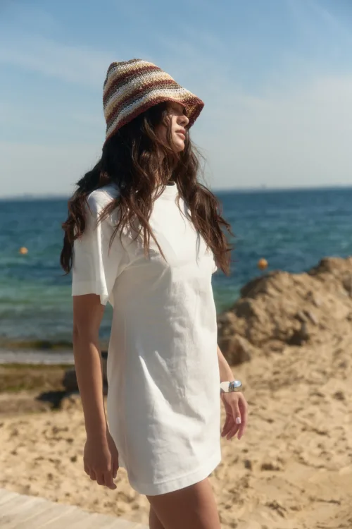 Woman at the beach wearing a striped hat and white dress, looking toward the sea with rocky shore in the background.