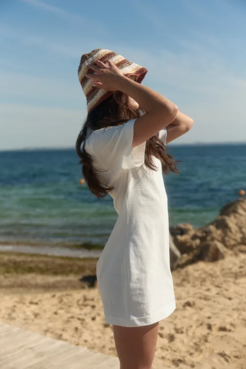 Woman at the beach wearing a striped knit hat and white dress, hands lifting the hat to shield her face, ocean in the background on a sunny day.