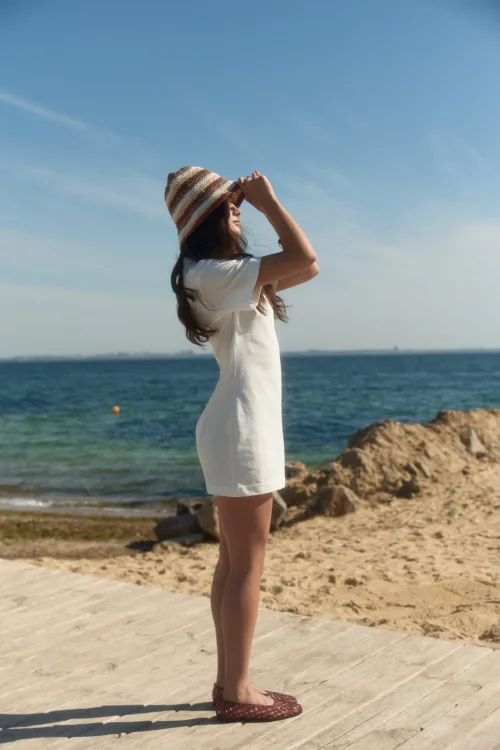 Young woman standing on a wooden boardwalk by the sea, adjusting a striped sun hat and wearing a white dress.
