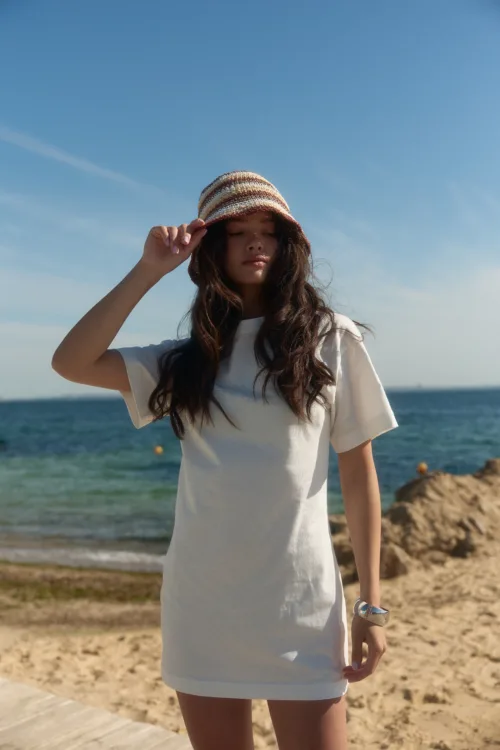 Woman standing on a sandy beach wearing a white dress and a striped sun hat, with the ocean in the background.