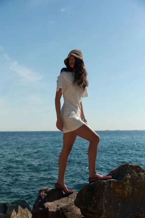 Person standing on rocky coastline by the sea, wearing a white dress and a woven hat, with blue sky above.