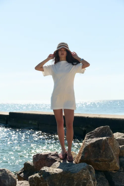 Woman standing on a rocky shoreline by the ocean on a sunny day, wearing a white dress and striped bucket hat.