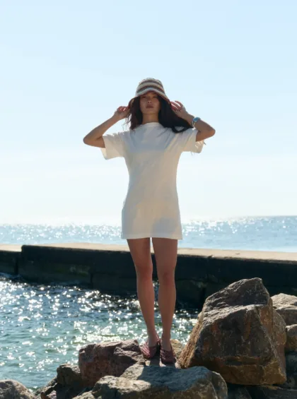 Woman standing on a rocky shoreline by the ocean on a sunny day, wearing a white dress and striped bucket hat.