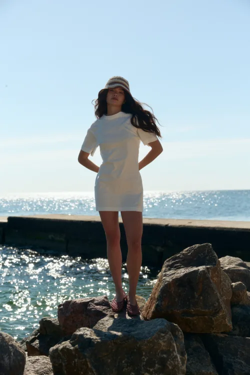 Woman in a white dress and striped hat standing on rocks at the seaside under a clear blue sky.
