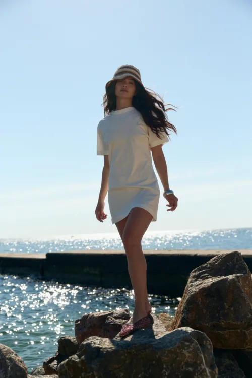 Woman standing on sunlit rocks by the sea, wearing a white dress and striped hat, with water sparkling in the background.