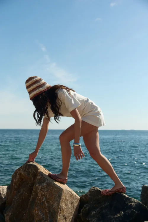 Person balancing on rocky shoreline by the sea, wearing a striped sunhat and white outfit.