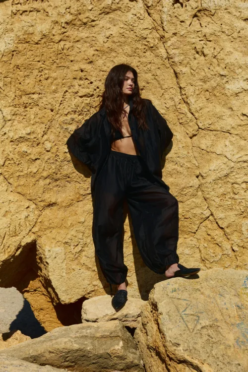 Woman with long dark hair sits on a sunlit beige rock wall wearing a loose black sheer outfit, looking to the side.