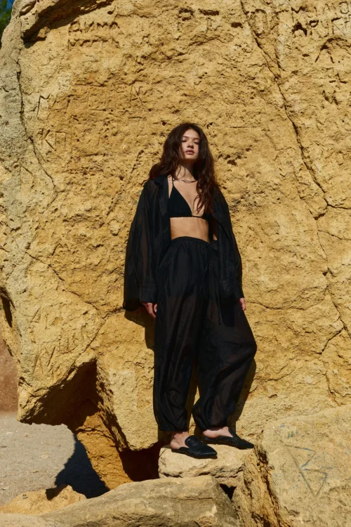 Young woman wearing a black bikini and sheer black cover-up stands against a large beige rock wall.