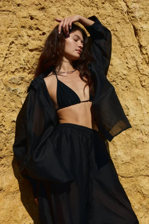 Woman in a black bikini top and sheer black cover-up posing with eyes closed against a rocky tan wall.