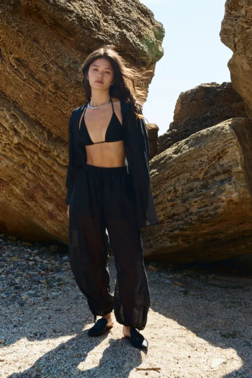 Woman in a black bikini top and sheer black pants stands between large rocky cliff formations on a pebbled beach, facing the camera.