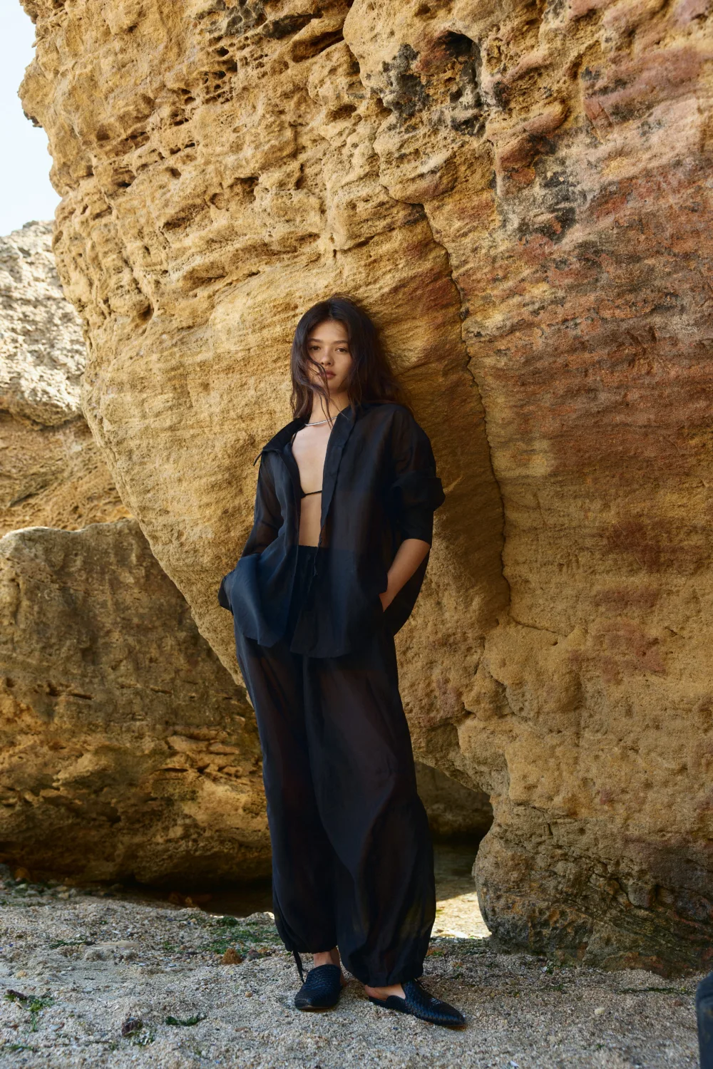 Woman in a black sheer outfit leaning against a textured rock cliff at the beach.