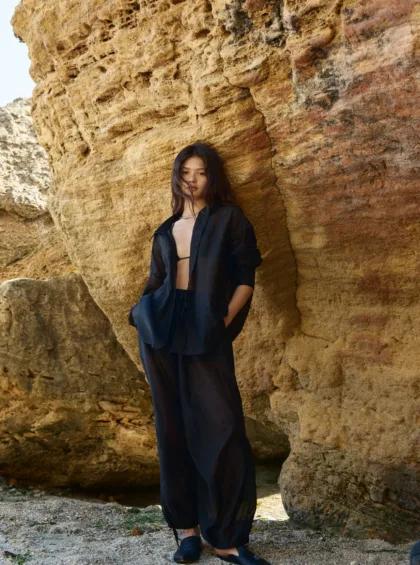 Woman in a black sheer outfit leaning against a textured rock cliff at the beach.