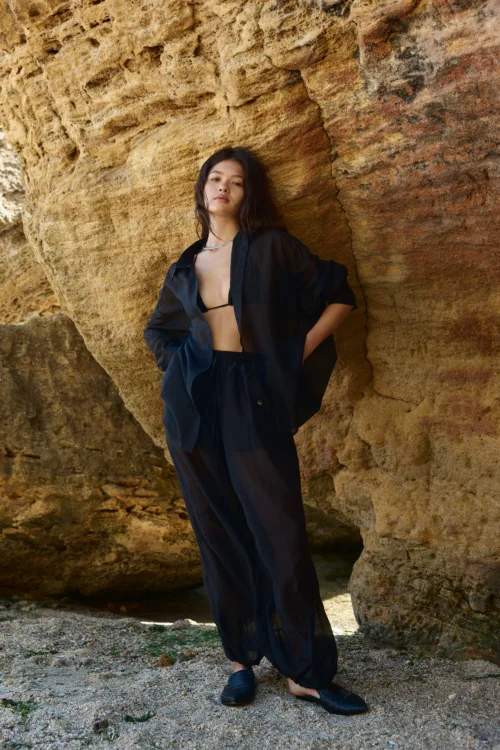 Woman in a black sheer outfit stands against a textured rocky wall outdoors, looking at the camera.