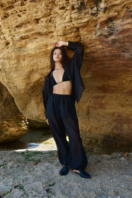 Woman in a black bikini and loose black outfit posing against a textured rock wall outdoors by the shore, with one arm raised above her head.