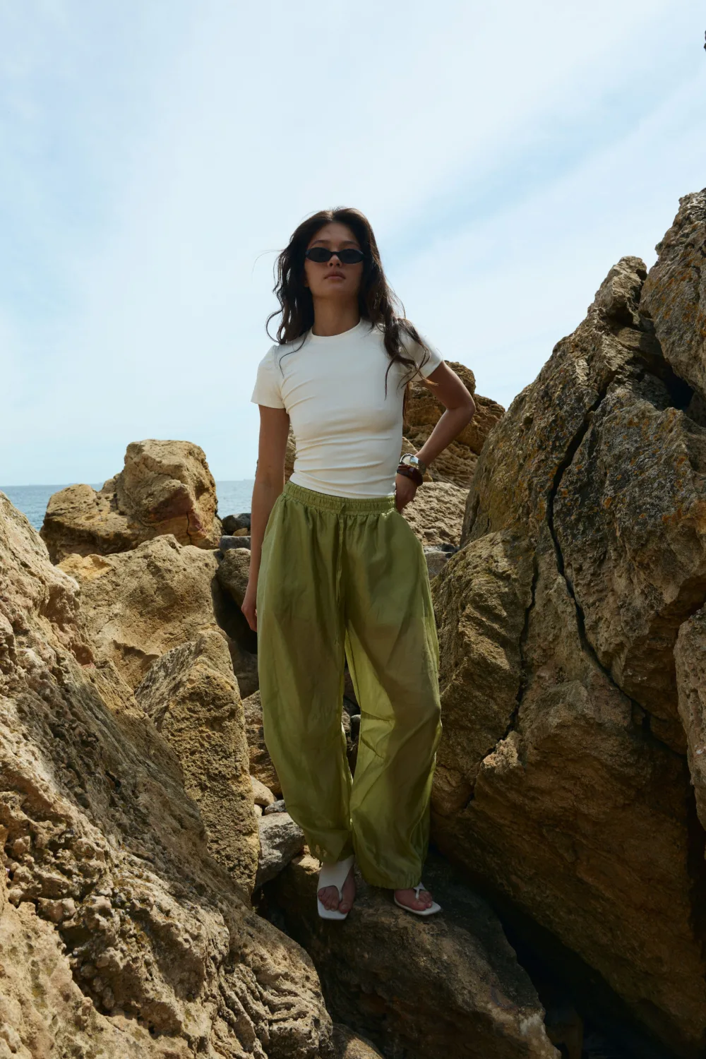 Woman standing on rugged coastal rocks, wearing a white T-shirt, green loose pants, and sunglasses with the sea in the background.
