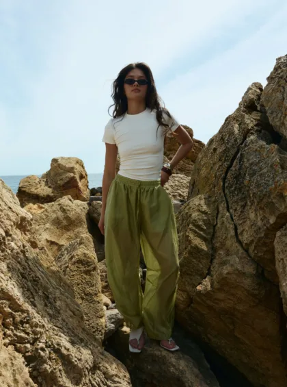 Woman standing on rugged coastal rocks, wearing a white T-shirt, green loose pants, and sunglasses with the sea in the background.