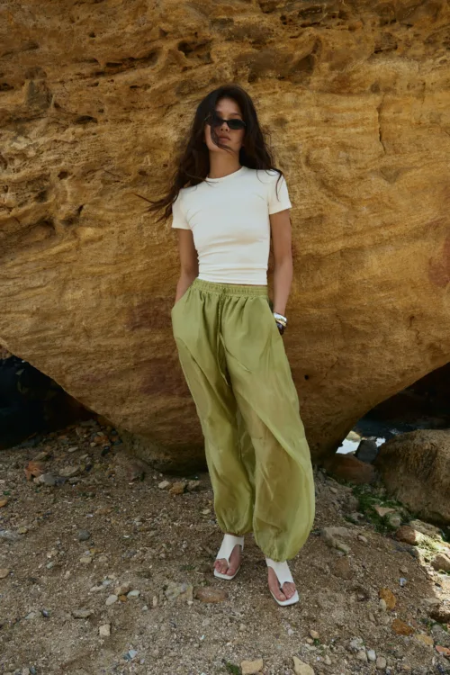Woman standing in front of a textured brown rock wall, wearing a white short-sleeve top, loose green pants, white sandals, and dark sunglasses, with hair blown by the wind.