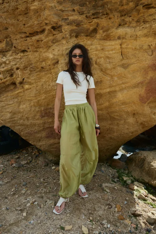 Woman posing outdoors against a large rocky wall, wearing a white short-sleeve top, olive green pants, and sunglasses.