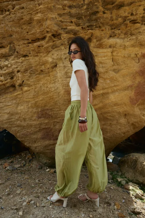 Woman in a white short-sleeve top and olive wide-leg pants, wearing white heeled sandals, stands beside a textured brown rock wall.