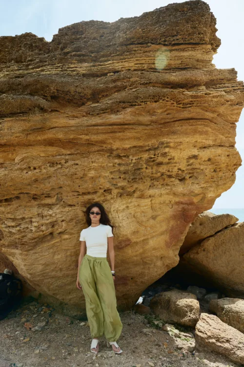 Woman in a white t-shirt and olive-green pants stands at the base of a large layered rock formation by a rocky beach, wearing sunglasses.