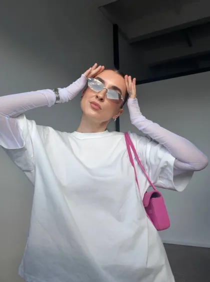Woman posing in a white oversized shirt with lavender sleeves, wearing mirrored sunglasses and a pink purse, holding sides of her head.