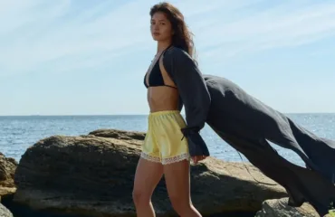 Woman standing on rocky shore by the sea, wearing yellow shorts with lace trim and a black bikini top, a dark flowing cover-up billowing behind her in the wind.