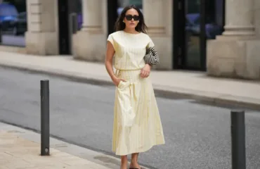 Woman in a pale yellow striped midi dress walking on an urban sidewalk, wearing sunglasses and holding a black-and-white striped clutch bag.