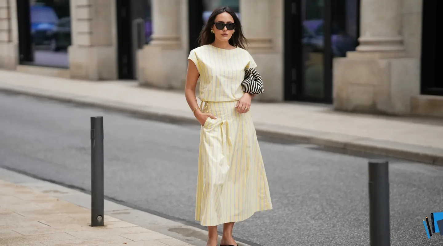 Сукні жіночі на літо: актуальні фасони - Woman in a pale yellow striped midi dress walking on an urban sidewalk, wearing sunglasses and holding a black-and-white striped clutch bag.