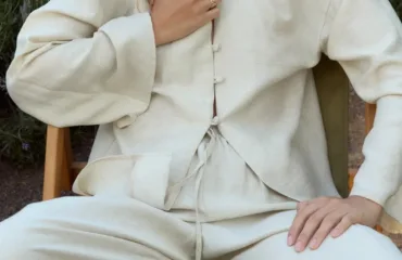 Person seated outdoors wearing a cream linen suit with a buttoned jacket and tied waist.