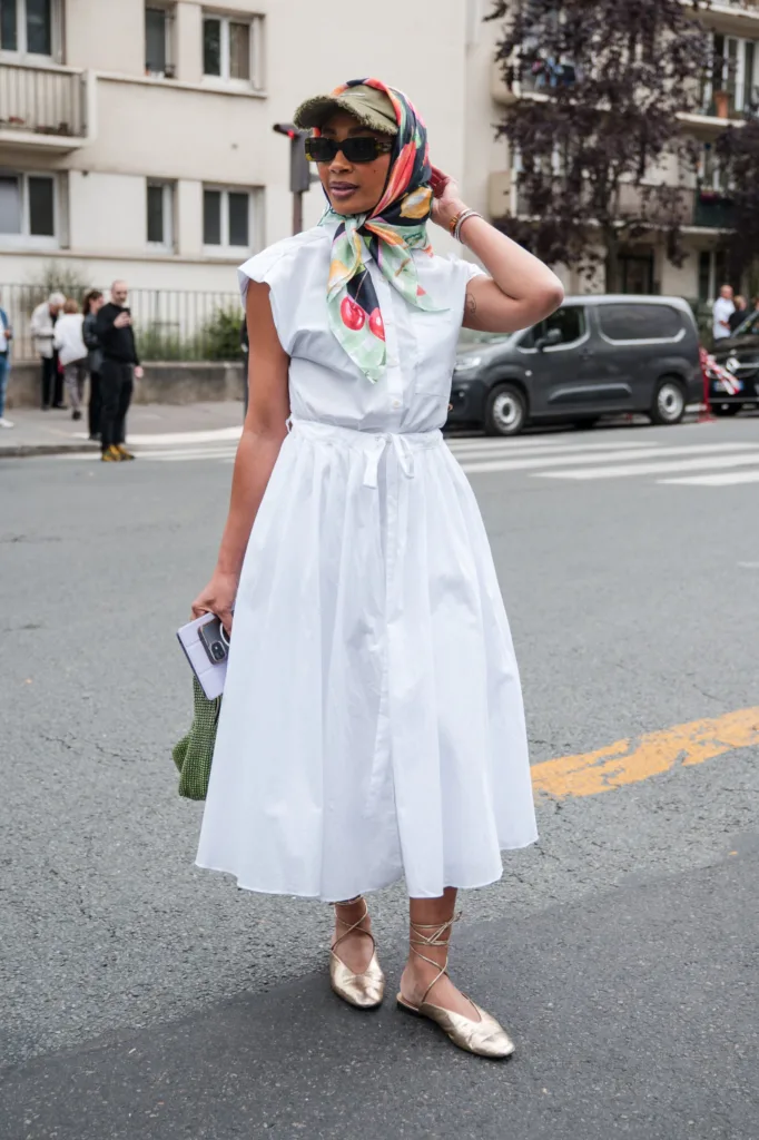 Жіночі аксесуари для літніх образів | Romanova - Woman wearing a white midi dress with a tied waist, colorful headscarf, sunglasses, and beige sandals on a city street.