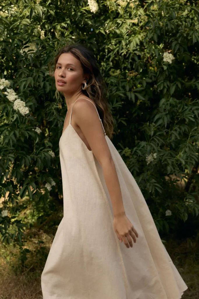 Сукні жіночі на літо: актуальні фасони - Woman in a beige sleeveless dress walks beside a hedge with white blossoms, looking toward the camera.