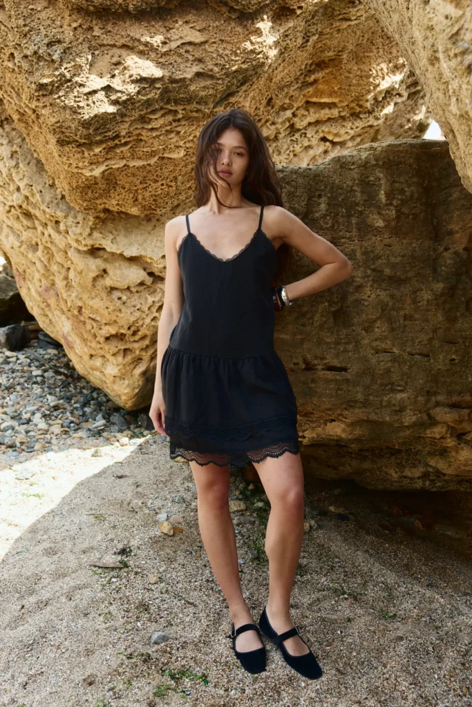 Сукні жіночі на літо: актуальні фасони - Woman in a black lace-trim dress standing against a rocky beach wall, sunlit stones around her.