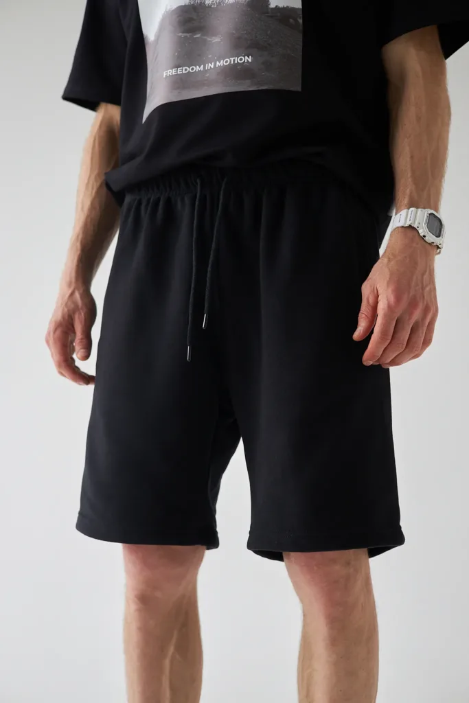 Шорти для всієї родини: моделі на літо - Man wearing a black graphic T-shirt and black drawstring shorts, with a white digital watch on his left wrist.