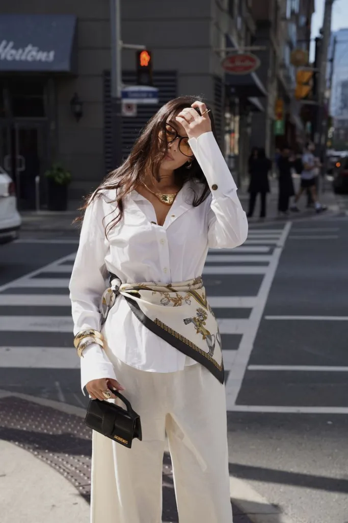 Жіночі аксесуари для літніх образів | Romanova - Stylish woman in a white shirt and wide-leg pants, accessorized with a patterned scarf belt, sunglasses, and a small black handbag on a city crosswalk.