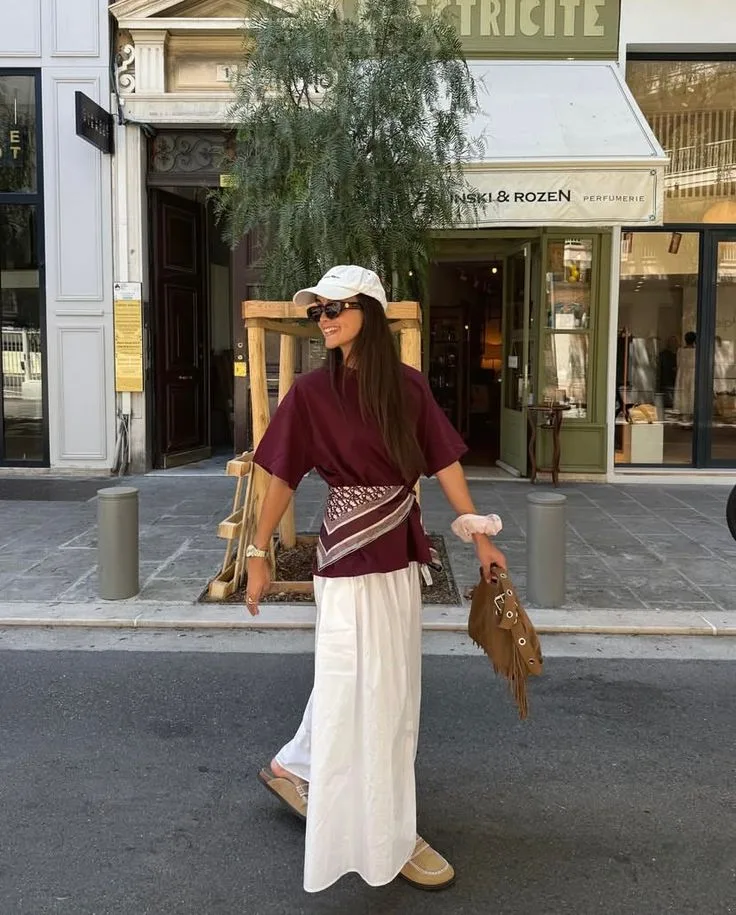 Жіночі аксесуари для літніх образів | Romanova - Woman in burgundy top and long white skirt walks on a city street, wearing a beige cap and sunglasses.
