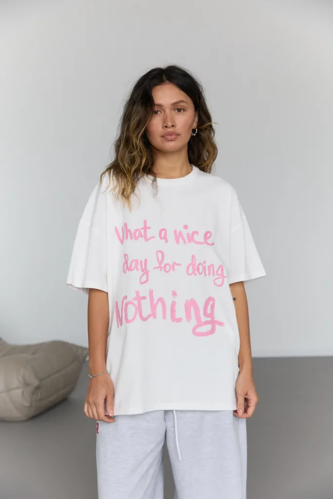 Жіночі футболки на літо: базові й oversize - Woman standing indoors wearing a white oversized T-shirt with pink text, looking at the camera.