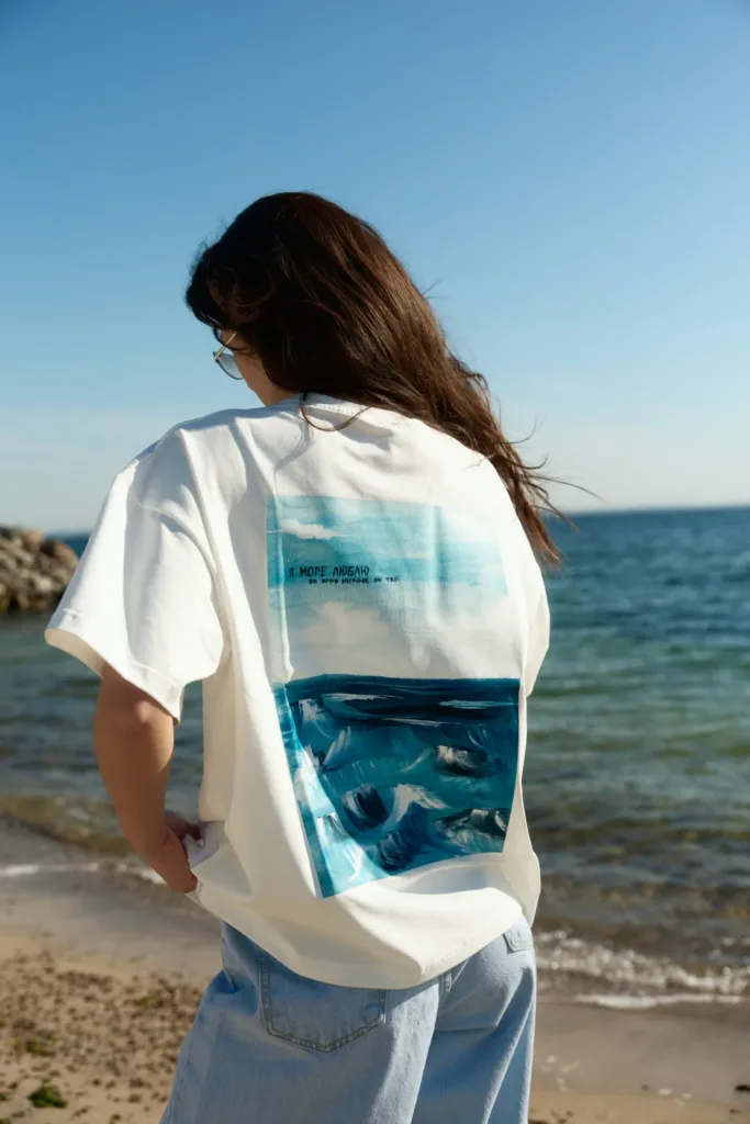 Жіночі футболки на літо: базові й oversize - Person standing on a sandy beach with their back to the camera, wearing a white T-shirt featuring a blue ocean graphic and light jeans, ocean in the background.