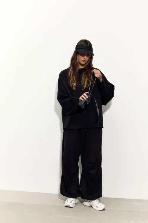 Person wearing an oversized black sweatshirt and black pants, white sneakers, and a black visor, holding a strap or bag.