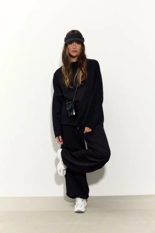 Woman in an all-black athleisure outfit with a black visor, white sneakers, and a camera hanging from a strap, posing against a light wall.