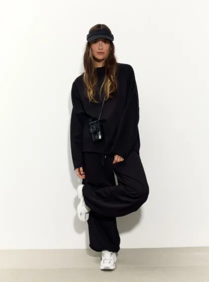 Woman in an all-black athleisure outfit with a black visor, white sneakers, and a camera hanging from a strap, posing against a light wall.