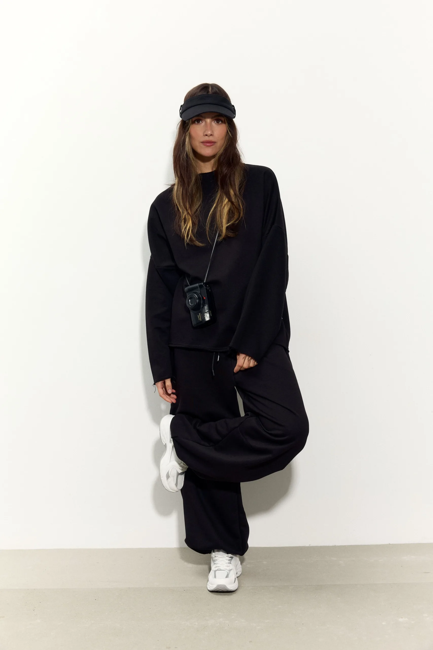 Жіночий чорний oversize костюм з м’якого футеру. Вільний силует і комфортна посадка для повсякденних та relaxed образів. Розмір xs/s, m/l - romanova4832_Original Костюм жіночий oversize чорний з футеру. Розмір xs/s, m/l - Woman in an all-black athleisure outfit with a black visor, white sneakers, and a camera hanging from a strap, posing against a light wall.