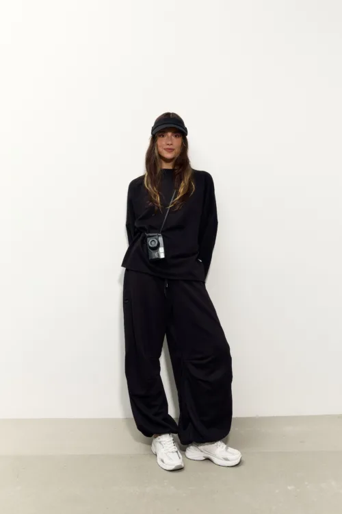 Woman in black athleisure with a crossbody camera, standing against a plain white wall wearing a black visor.