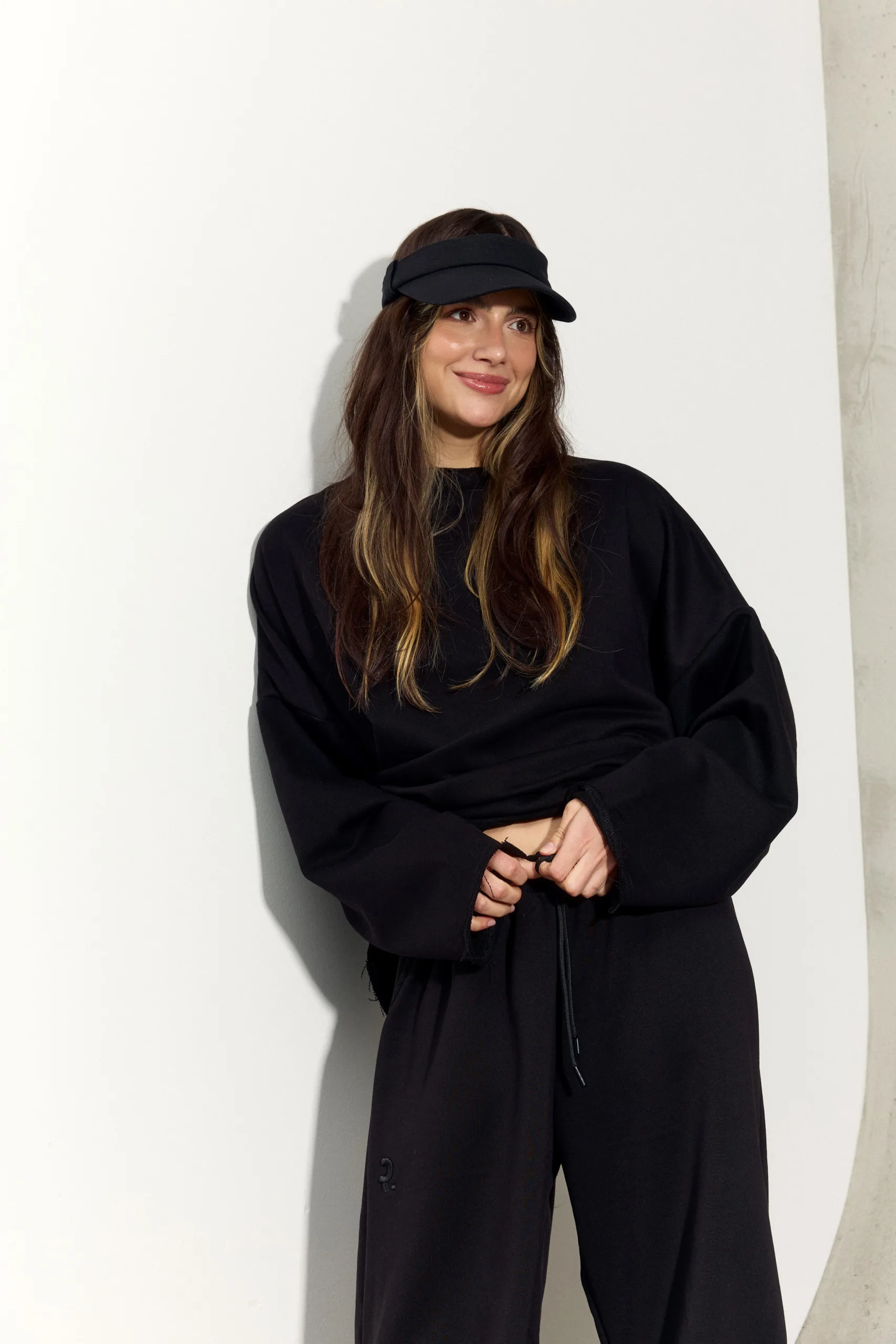 Жіночий чорний oversize костюм з м’якого футеру. Вільний силует і комфортна посадка для повсякденних та relaxed образів. Розмір xs/s, m/l - romanova4860_Original Костюм жіночий oversize чорний з футеру. Розмір xs/s, m/l - Woman with long brown hair wearing a black visor and all-black outfit, smiling against a pale wall.