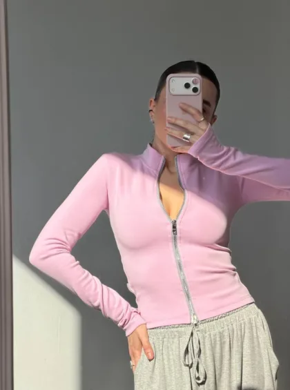 Person posing in a pink zip-up sweater and grey drawstring pants, taking a mirror selfie with a pink phone case obscuring their face.