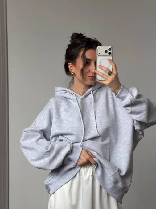 Худі жіноче сіре на блискавці з утепленням фліс - Woman in a light gray hoodie posing for a mirror selfie, hand in the pocket, wearing gold hoop earrings and white satin pants