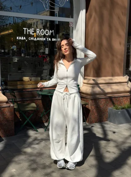 Woman in a white zip-front top and wide white pants poses outside a cafe named THE ROOM, eyes closed in sunlight.